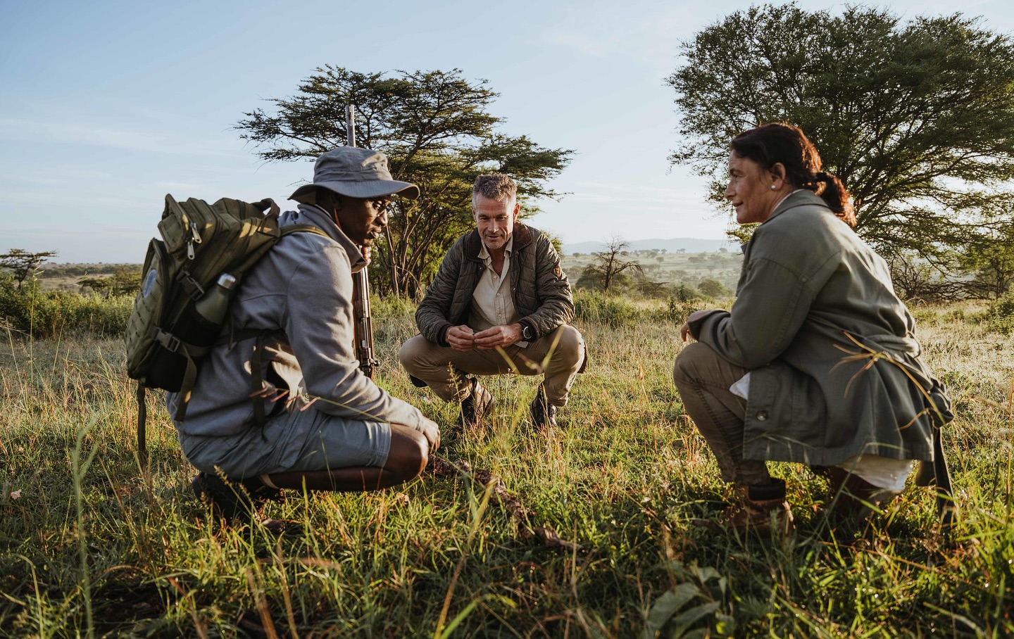 Three people in the wildlife crouch in a grassy expanse, discussing observations against a backdrop of trees and a bright sky.