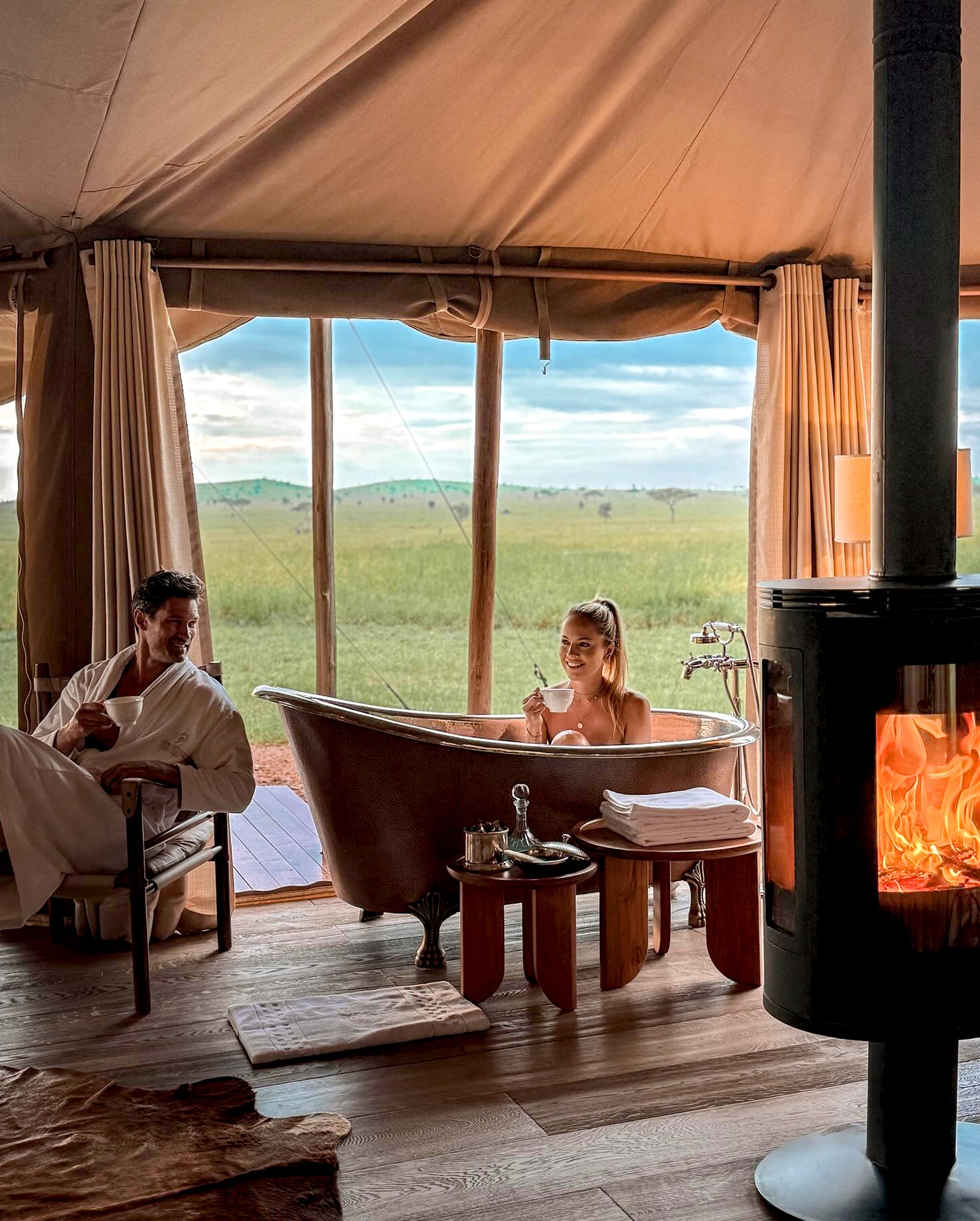 A cozy tent interior with a woman relaxing in a bathtub and a man sitting nearby, complemented by a warm fireplace and scenic views.